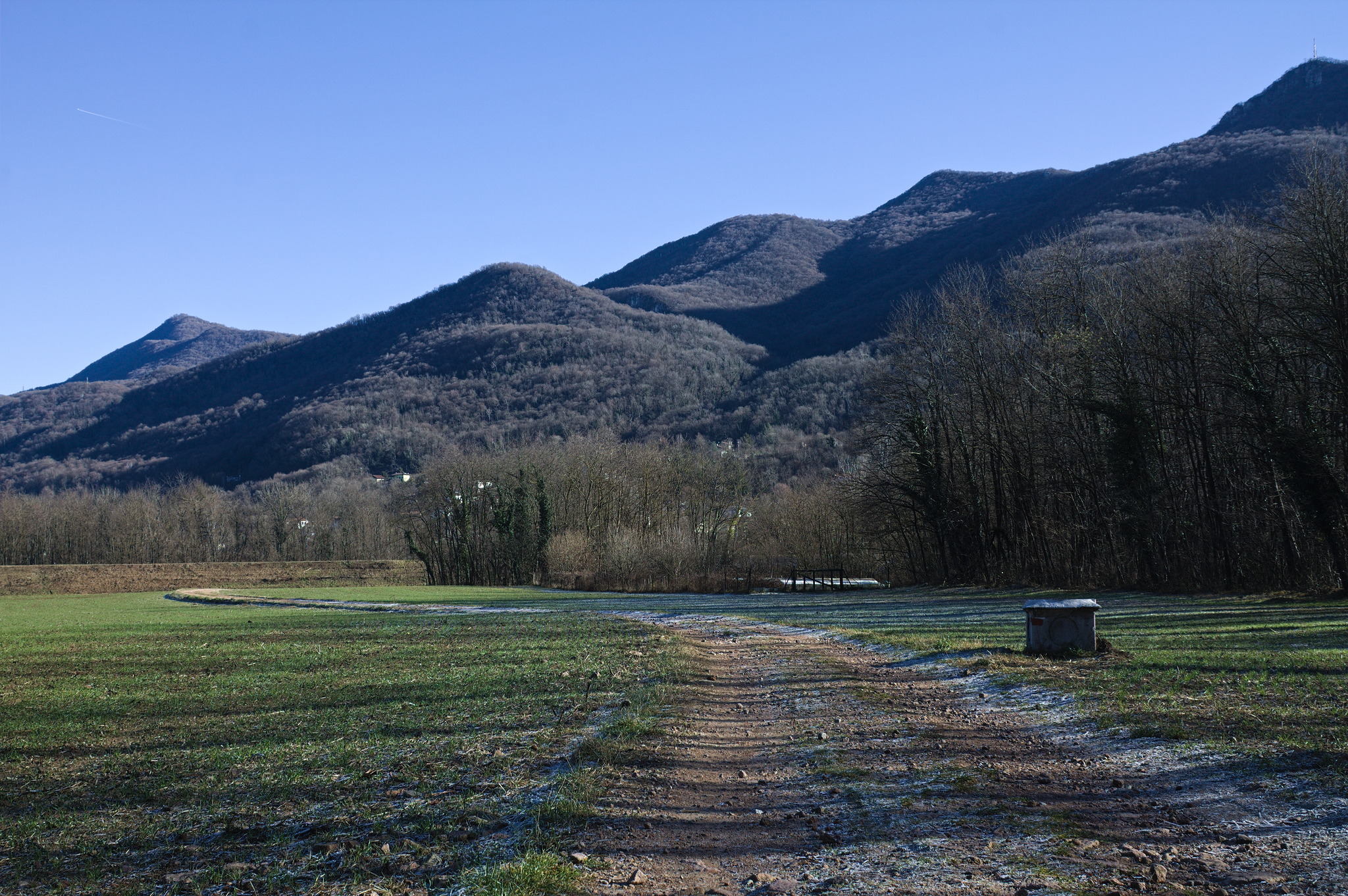 A wide and flat unpaved track passing through a flat grassy
area with trees to the sides and rolling hills in the background.