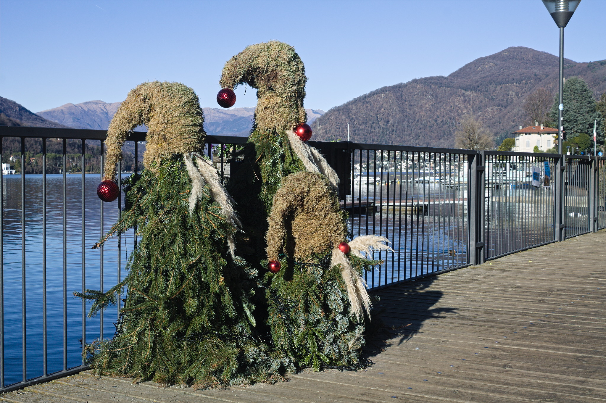 three gnome adjacent creatures made of branches of evergreen
trees, with a pointy hat made of moss, big white moustaches and
red Christmas tree balls at the end of the hat and as a nose.
They are wrapped in LED strings, and the lake can be seen in the
background.