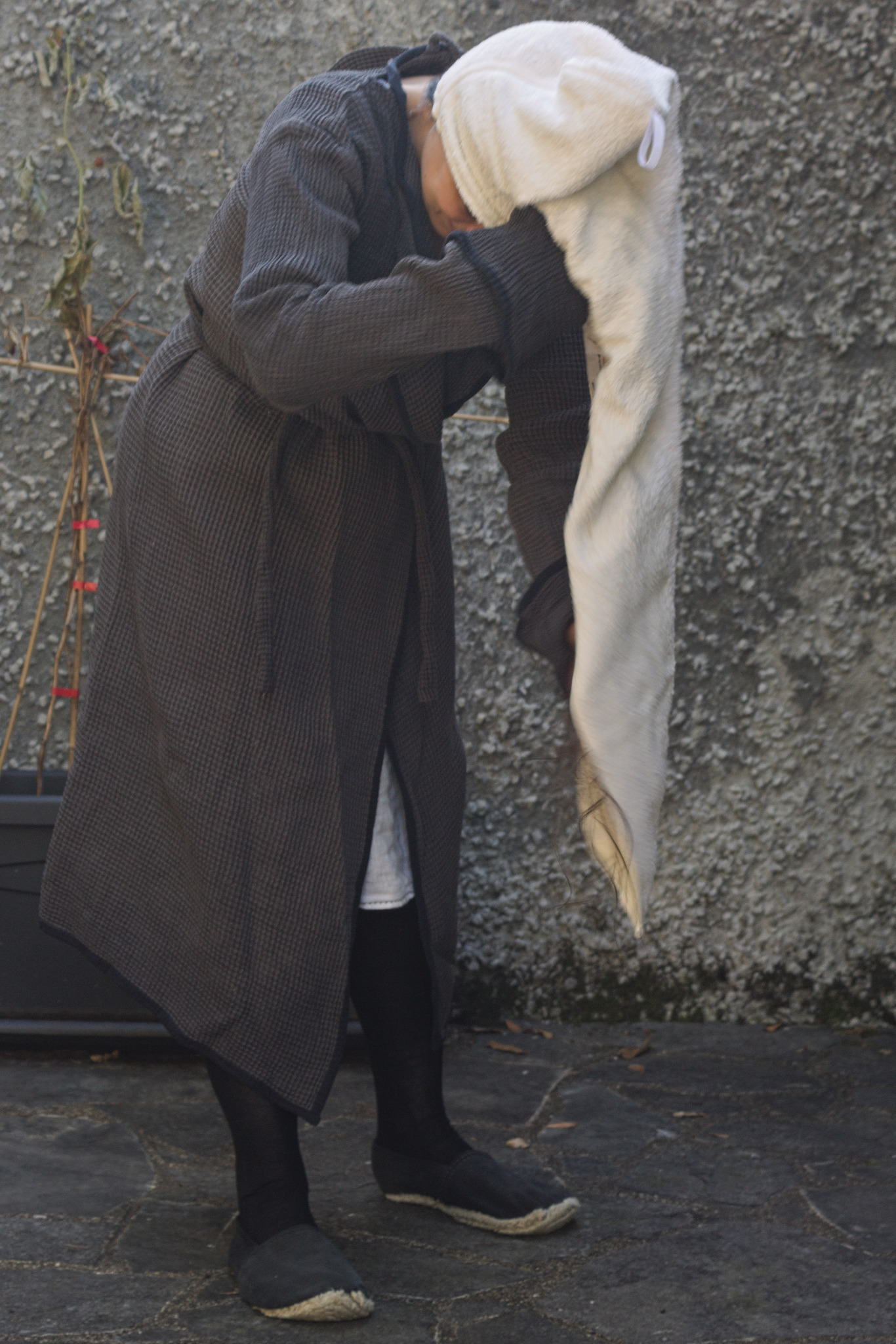 A woman in a bathrobe with her head tilted downwards, covered
by a towel thing that lies on the back of the head and has a long
tail hanging on the front, in the process of being wrapped around
the hairs and then brought over the top and back.