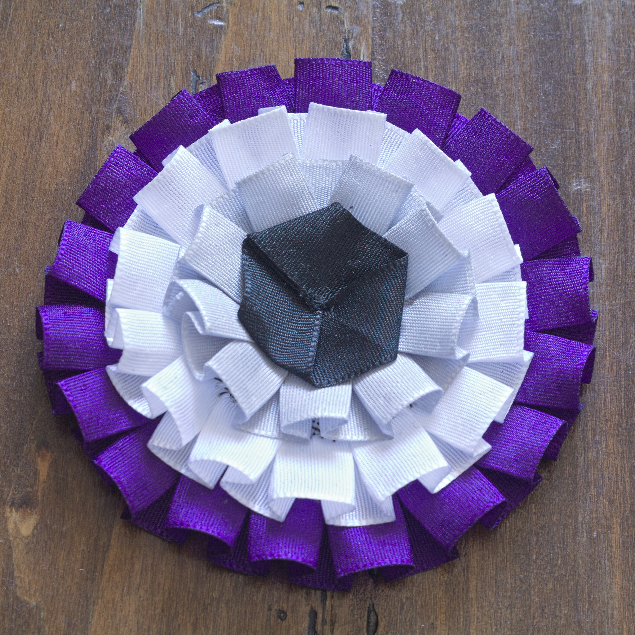 A cockade with three ribbons, starting with purple on the
outside, then white and grey, and an hexagonal shape made of black
ribbon at the centre. The ribbons are again box pleated.