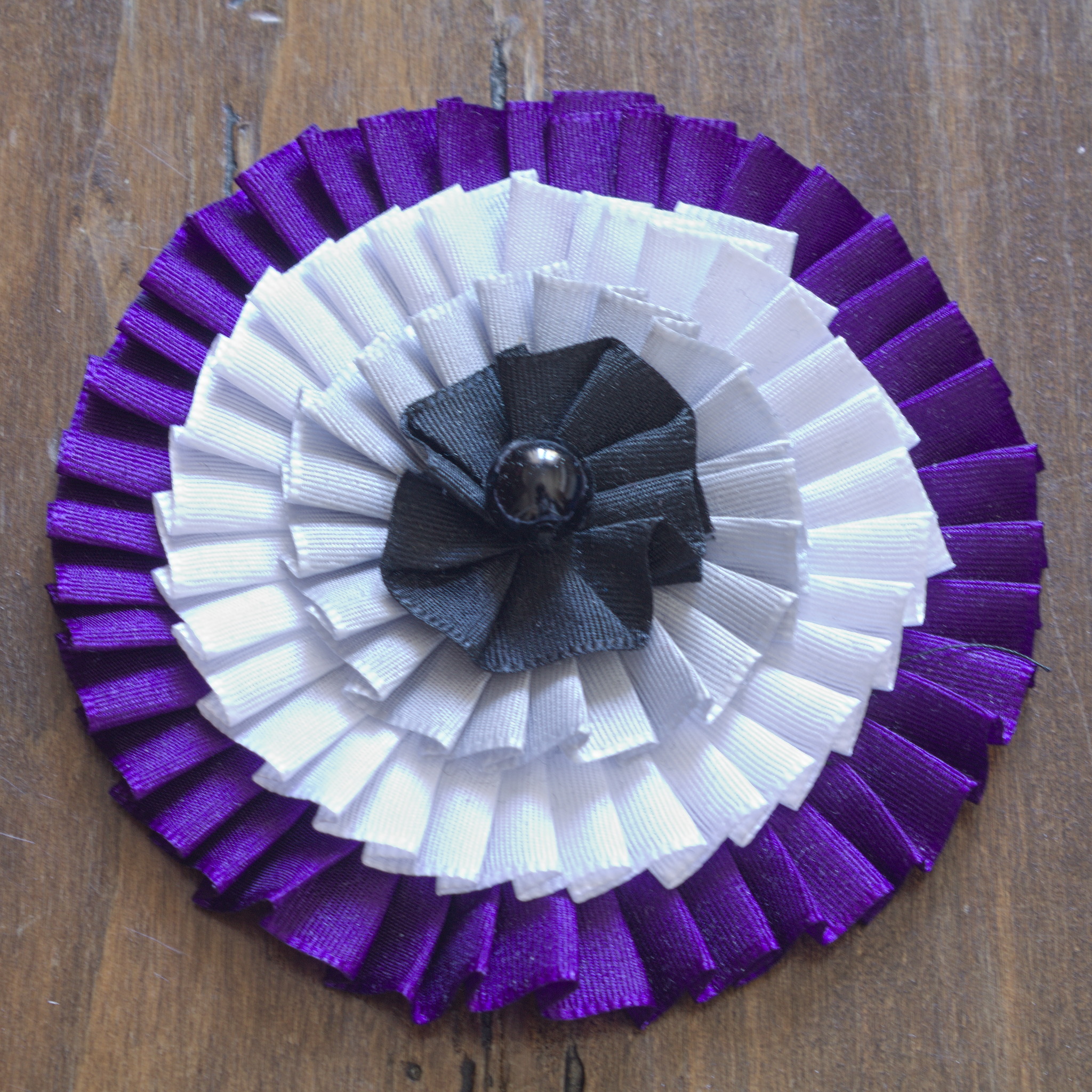 A big cockade with four ribbons, starting with purple on the
outside, and a black bead in the middle covering the centre of the
black ribbon. The pleating isn't the most regular, and the black
ribbon at the centre is almost more gathered than pleated.