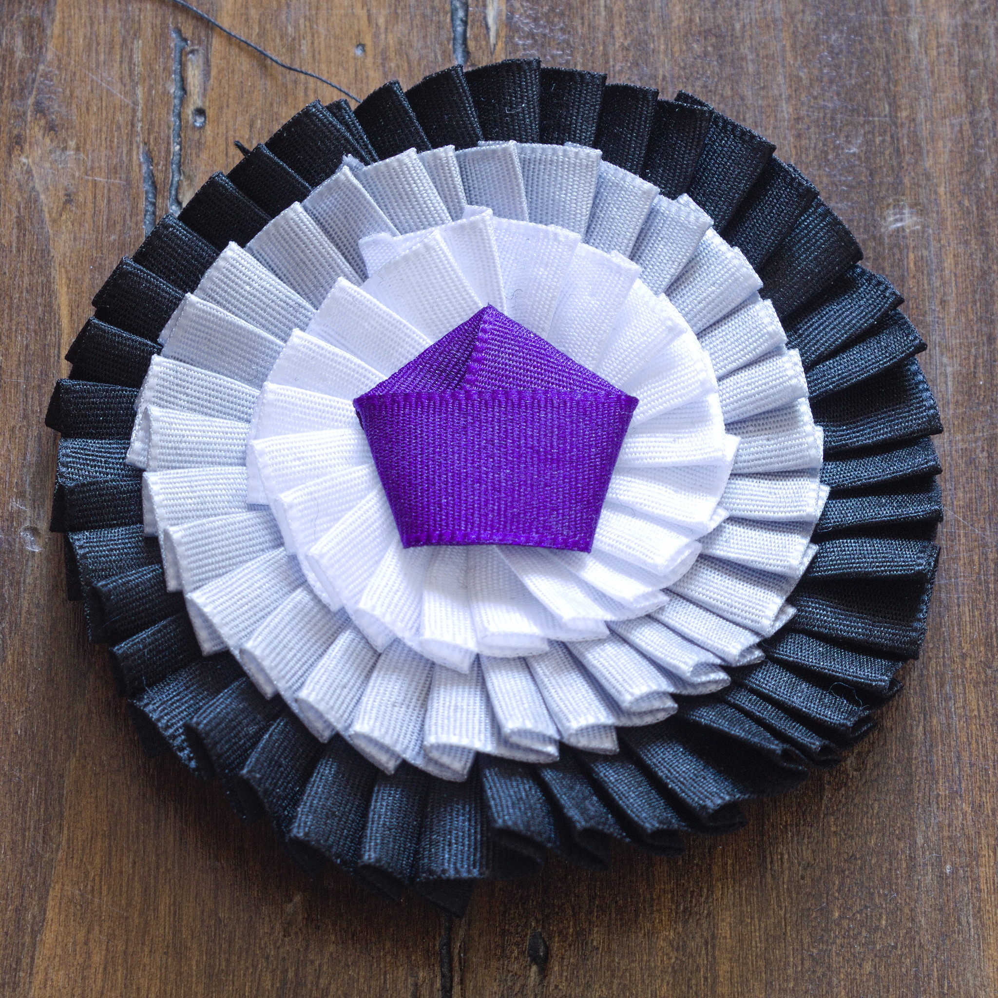 A cockade with three ribbons, starting with black on the
outside, then grey and white, and a flat pentagonal knot of purple
ribbon at the centre.