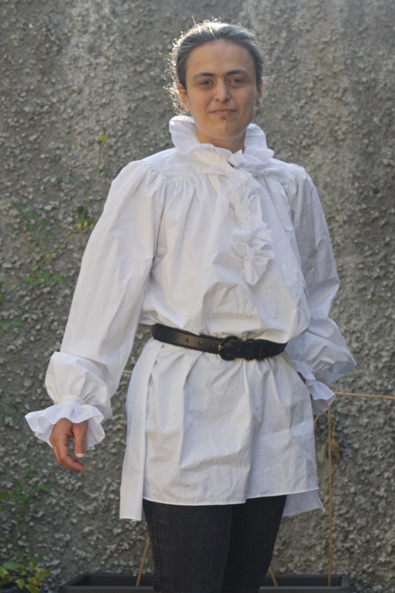A woman wearing an old-style white shirt with lots of fullness,
wide and long sleeves and ruffles at the collar that spread out
framing the neck, down the center front to underbust height,
covering the slit and at the cuffs, reaching to mid-hand. The
shirt is gathered at the waist with a belt, and worn over the
bottom garment to show that it reaches to mid tight.
Drama levels in the pose are pretty low.