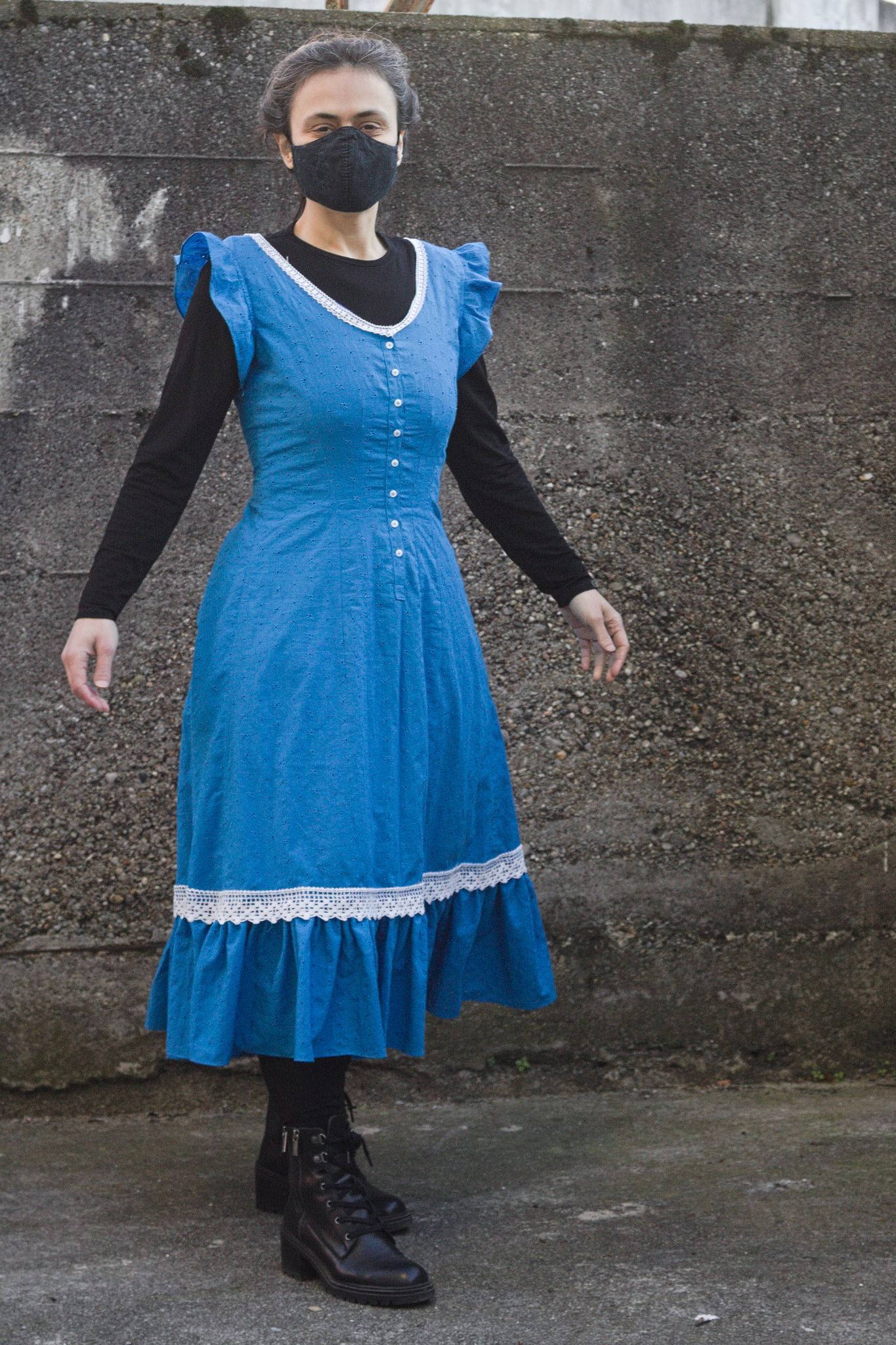 a person wearing a blue sleeveless fitted dress with
calf-length skirt; there are small ruffles on the armscyes and
the hem, and white lace on the collar and just above the hem
ruffle, and small white buttons on a partial placket down the
center front.
