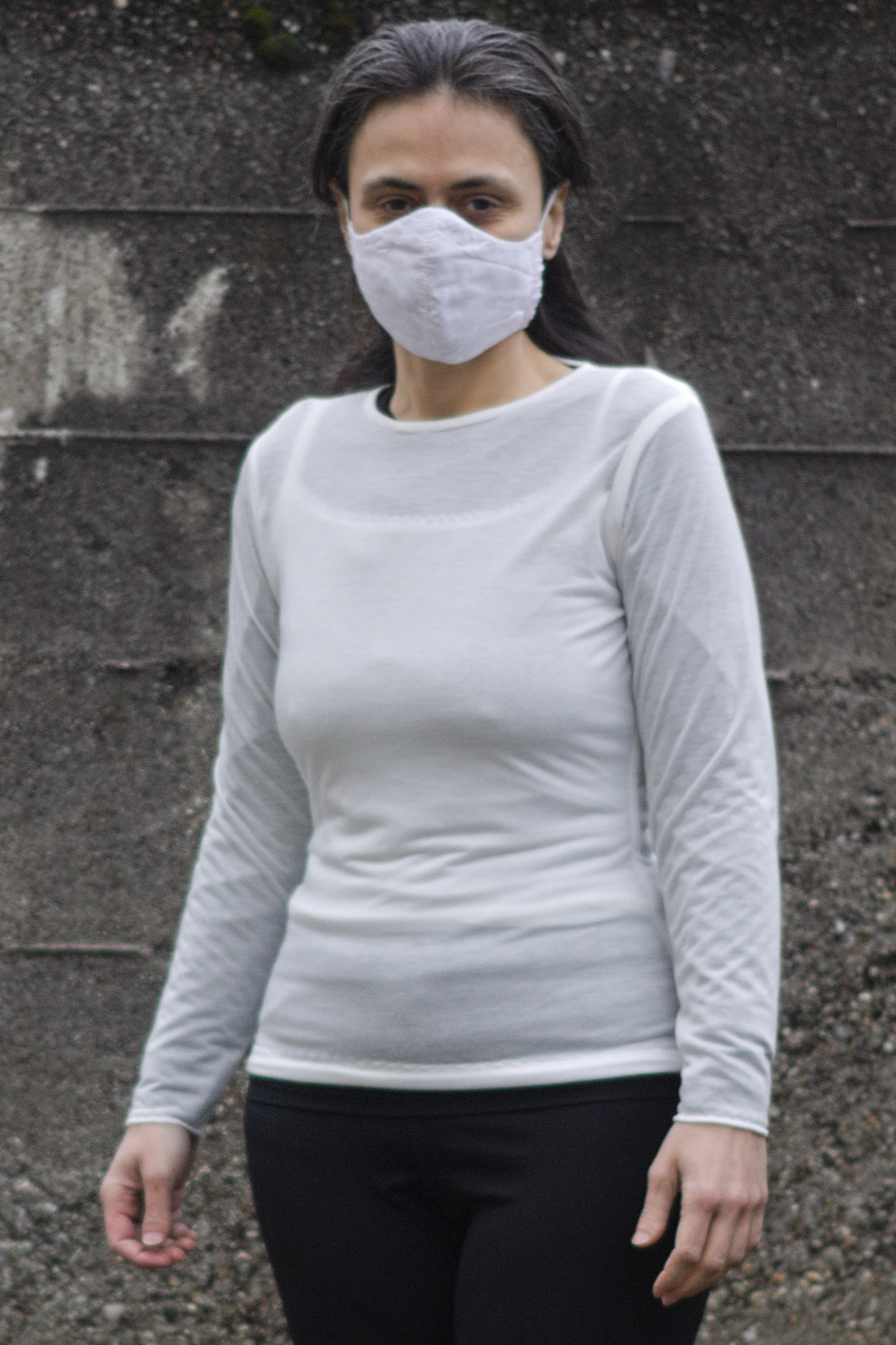 A woman wearing a simple, long sleeved, fitted white top; the fabric is somewhat transparent and the outline of a camisole can be seen.