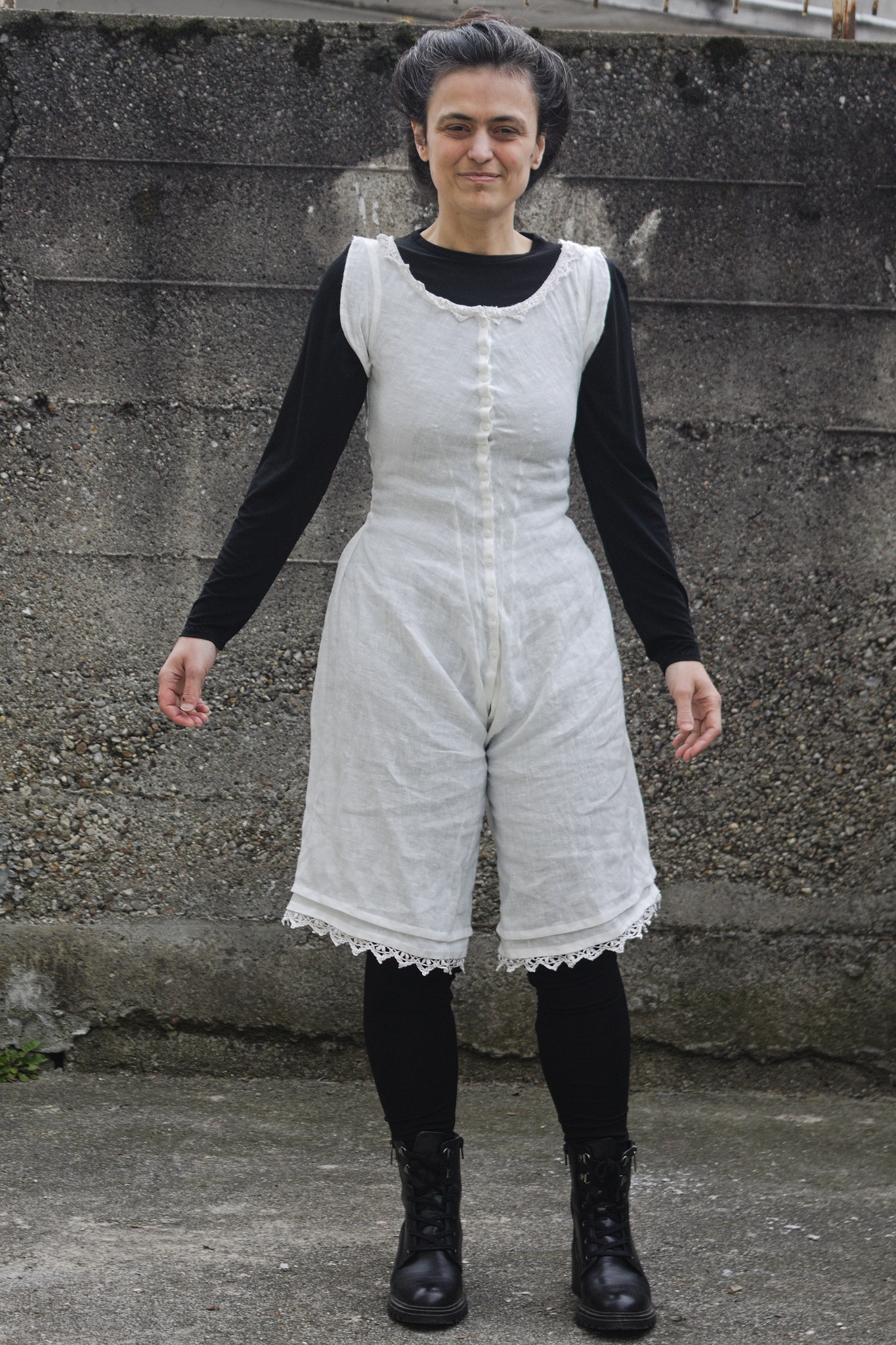 A woman wearing a white linen combination suite, with a very fitted top, small sleevelets that cover the armpits (to protect the next layers from sweat) and split drawers. The suite buttons up along the front (where it is a bit tight around the bust) and has a line of lace at the neckline and two tucks plus some lace at the legs.