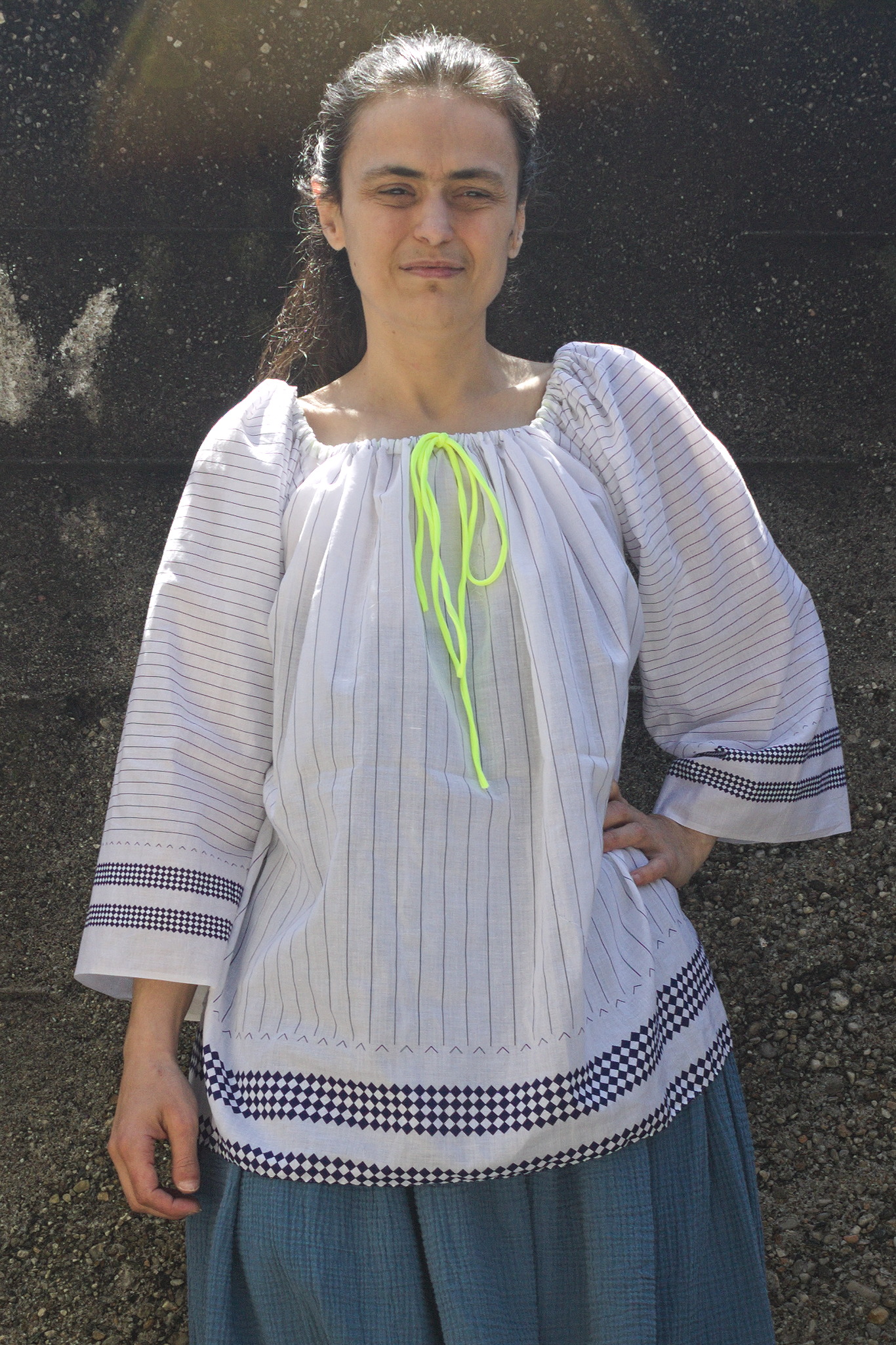 A woman wearing a top in white fabric with thin blue lines and two groups of blue lozenges near the hems. It has a square neck gathered by a yellow elastic, the blue lines are horizontal on the wide sleeves and vertical, and more spaced, on the body.