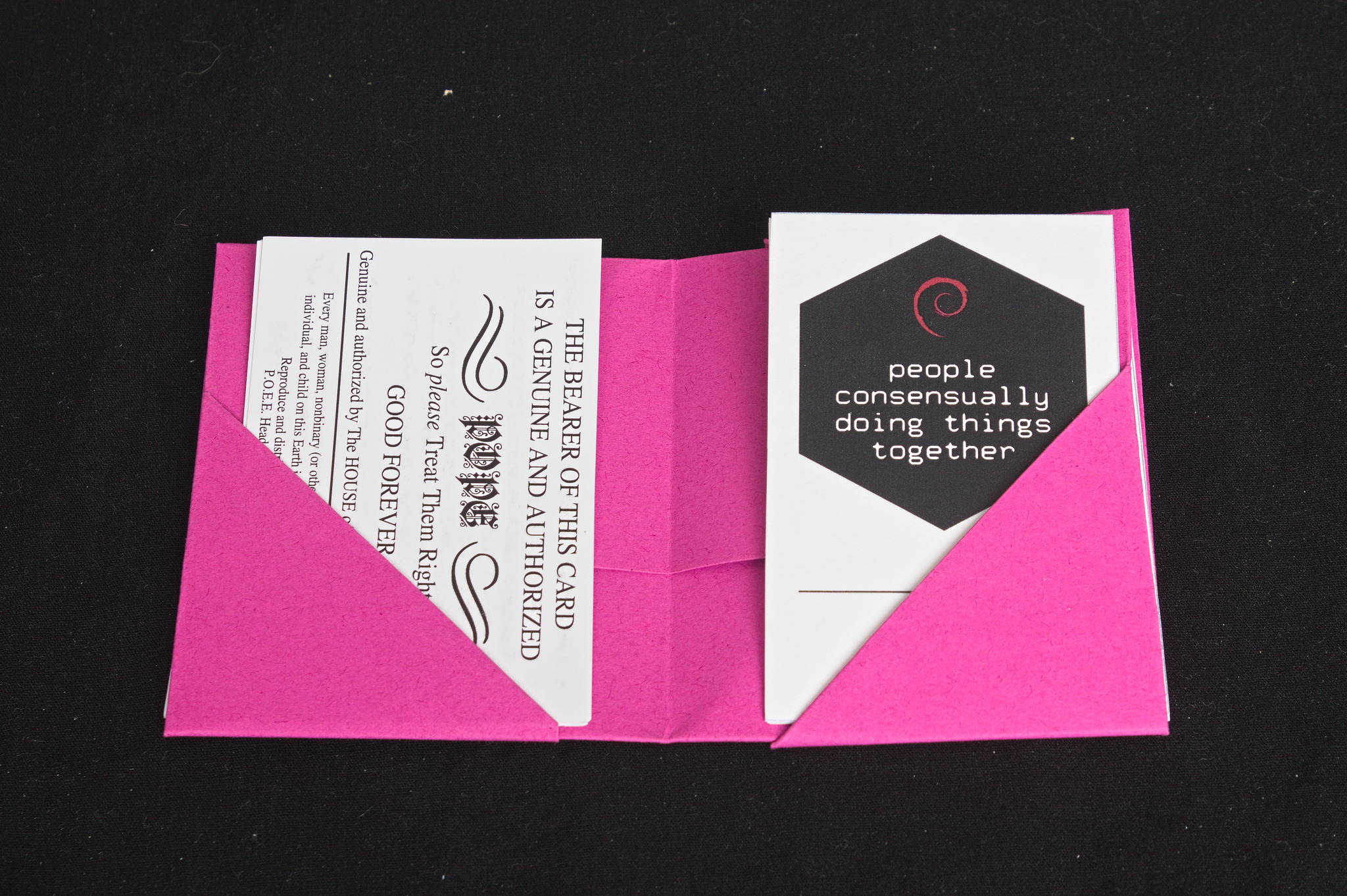 A pink origami holder for business cards, open, with two triangular pockets. The cards in it are pope cards and a Debian  consensually  name card.