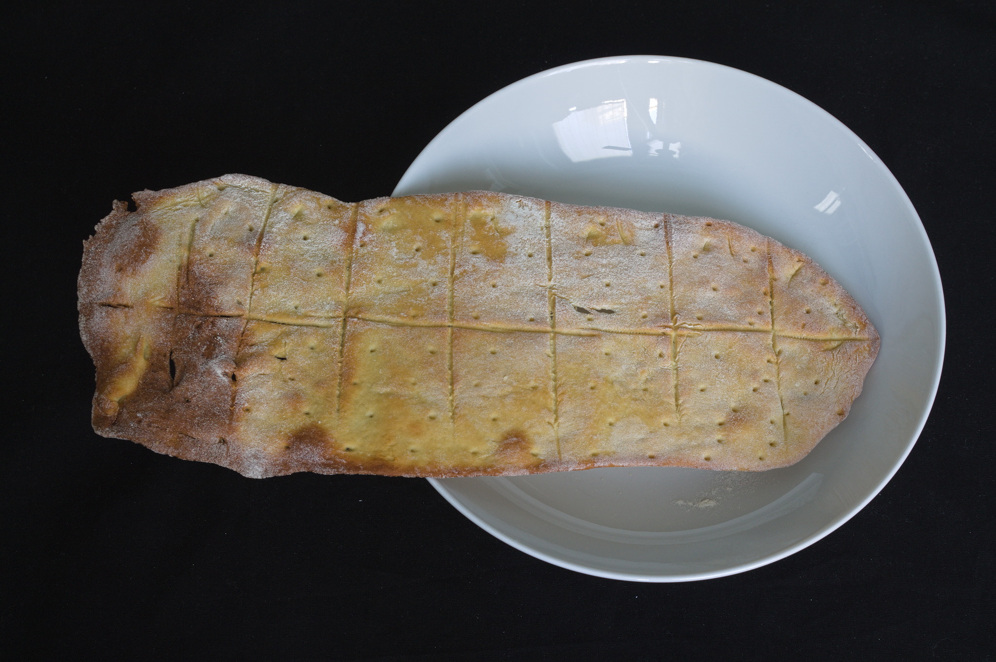 A flatbread, prescored into small portions, but still in one piece on top of a plate and overflowing to the side (it's about 10 cm   30 cm or so). A side is thin and more browned, the other side is a bit thicker and paler.