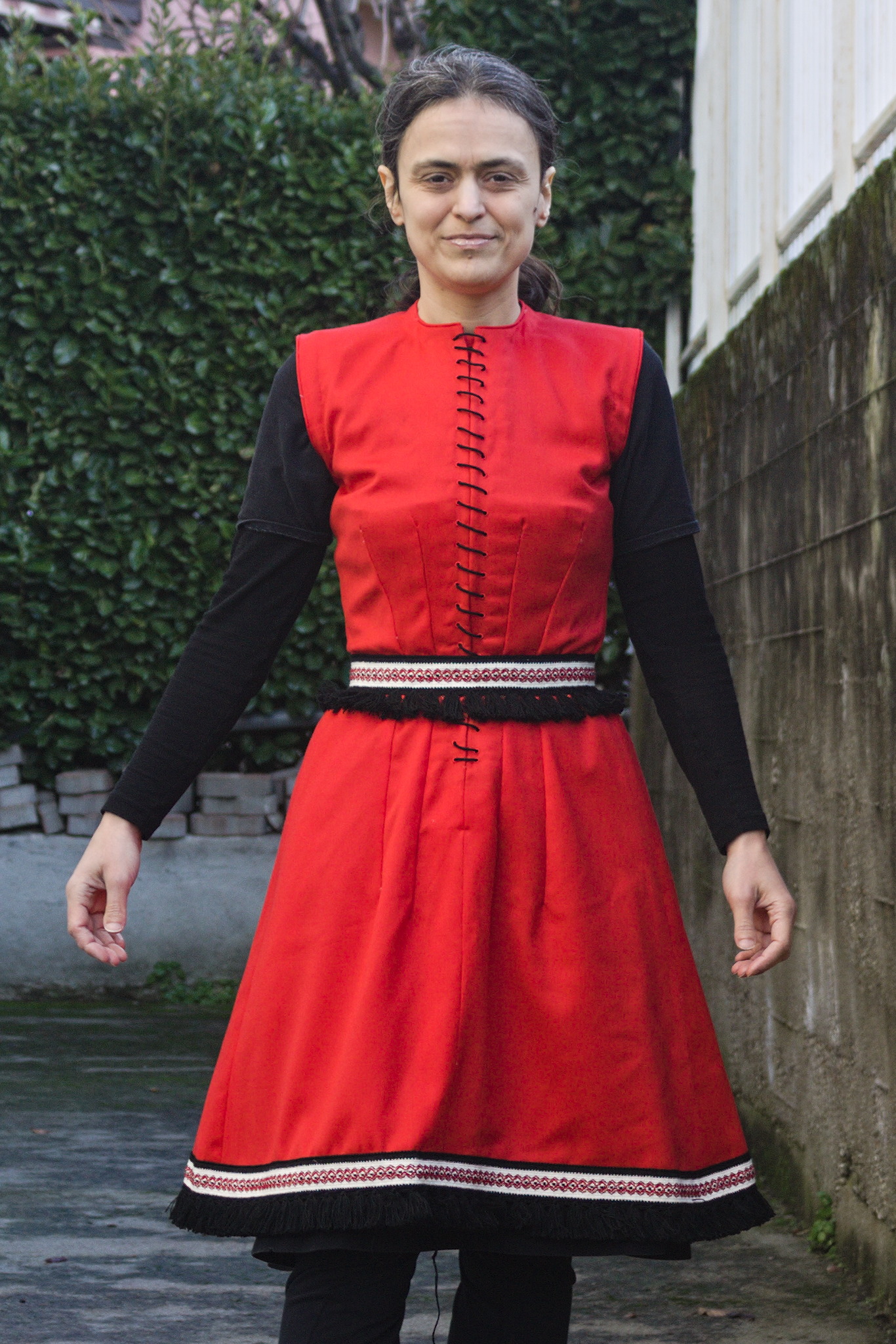 A woman wearing a red sleeveless dress; from the waist up it is
fitted, while the skirt flares out.
There is a white border with red embroidery and black fringe at
the hem and a belt of the same material at the waist.
