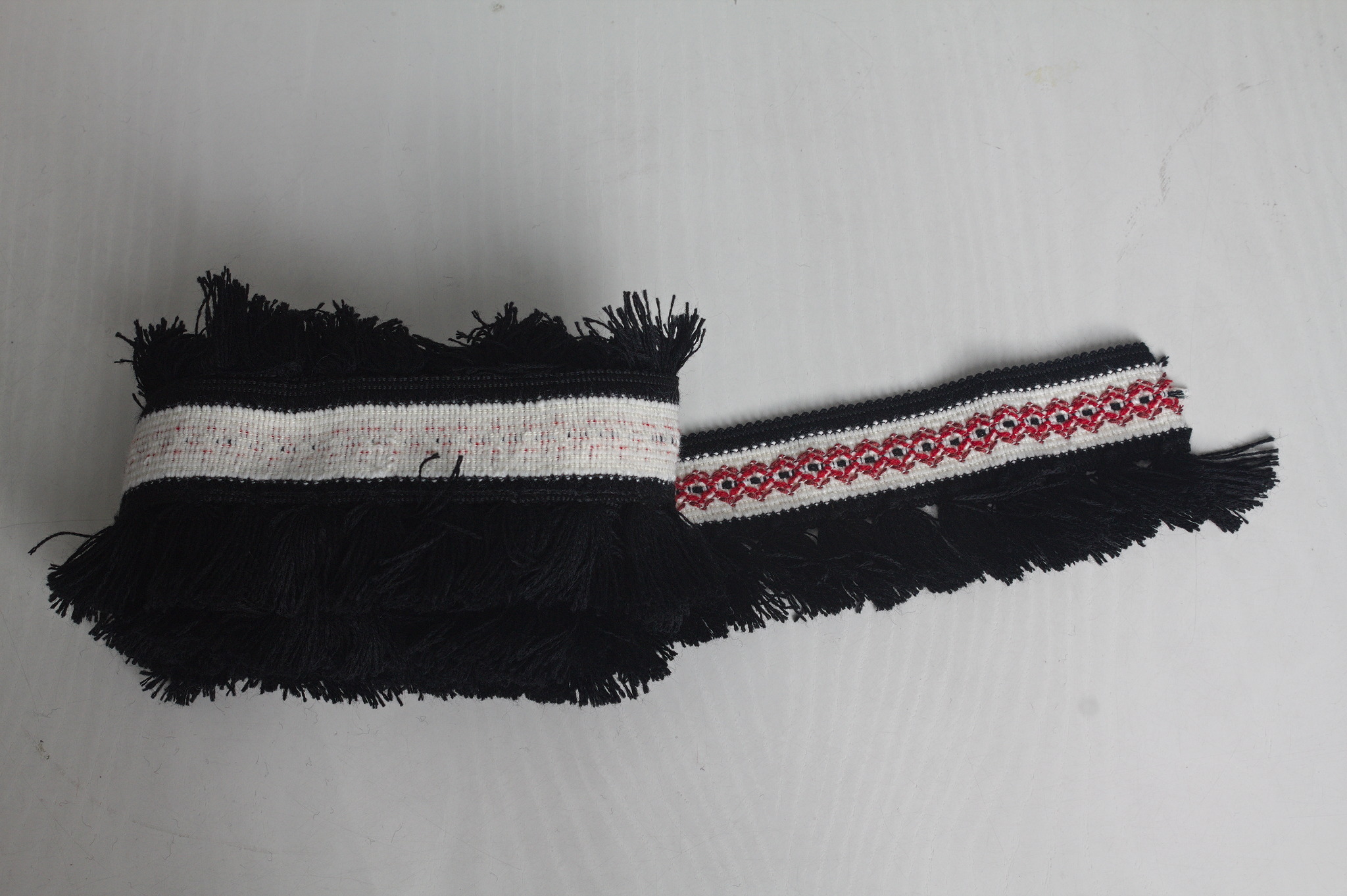 A few meters of wool-imitation fringe trim rolled up; the fringe is black and is attached to a white band with a line of lozenge outlines in red and brown.