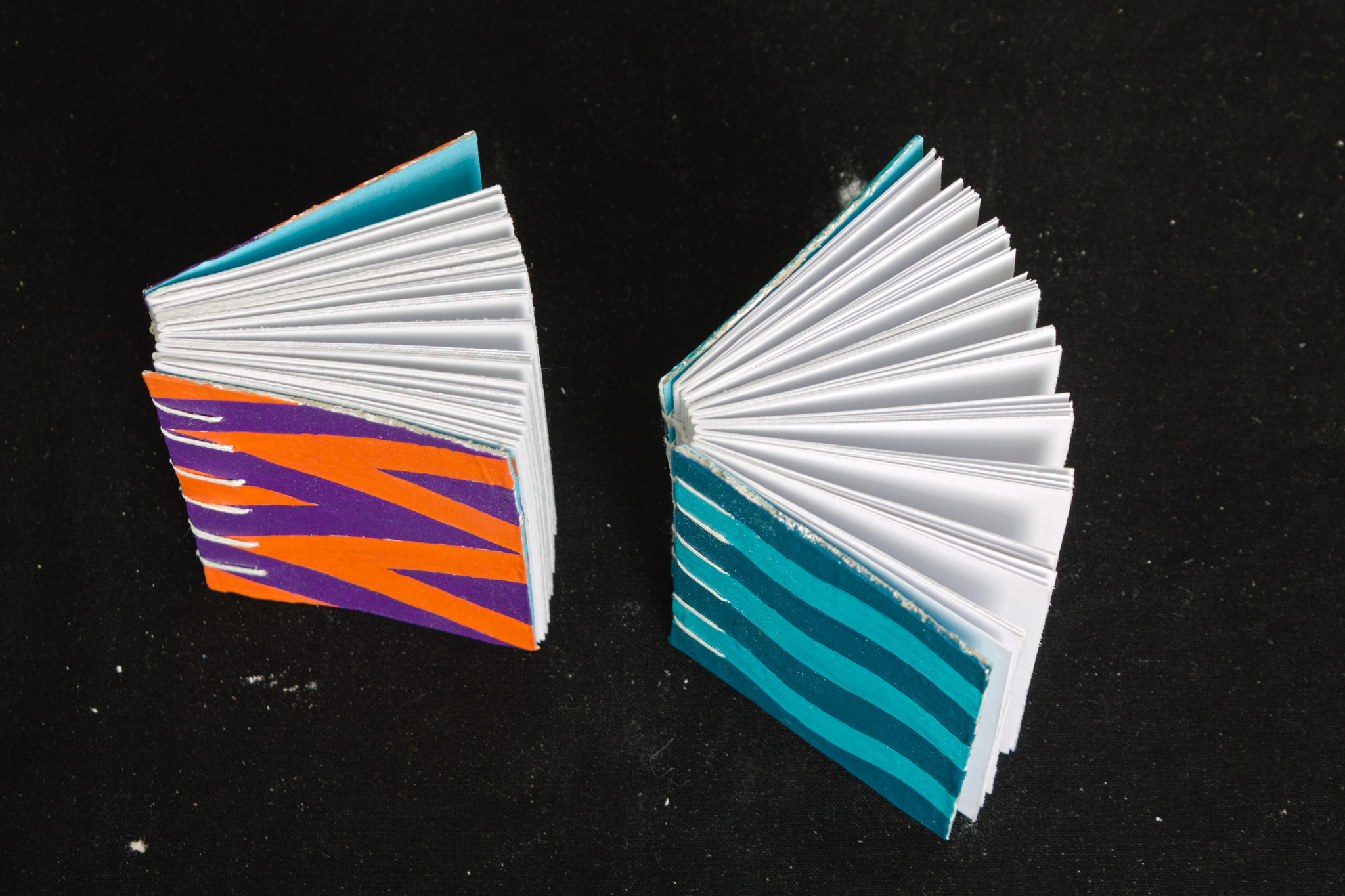 Two coptic bound small books, seen from the top with the pages
somewhat open. One has purple and orange triangles on the cover,
the other one waves in two shades of greenish blue.