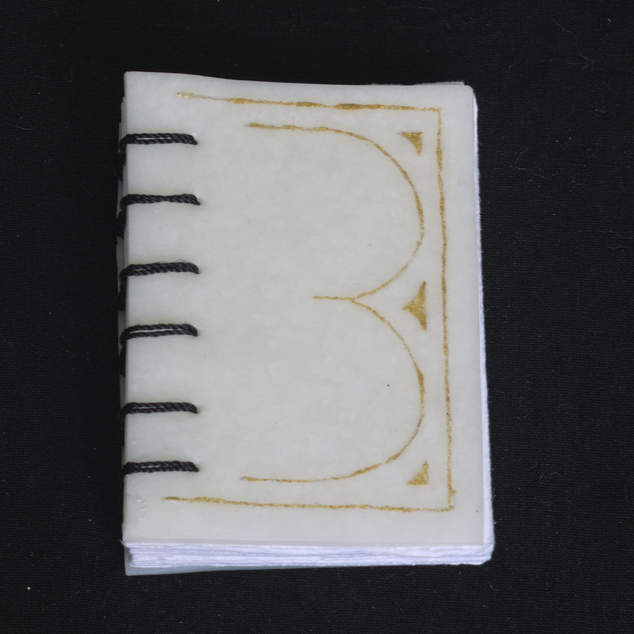 A coptic bound small book, seen from the front. The cover is
made of white cernit, with thin lines painted in gold acrylic to
form a sort of B shape.