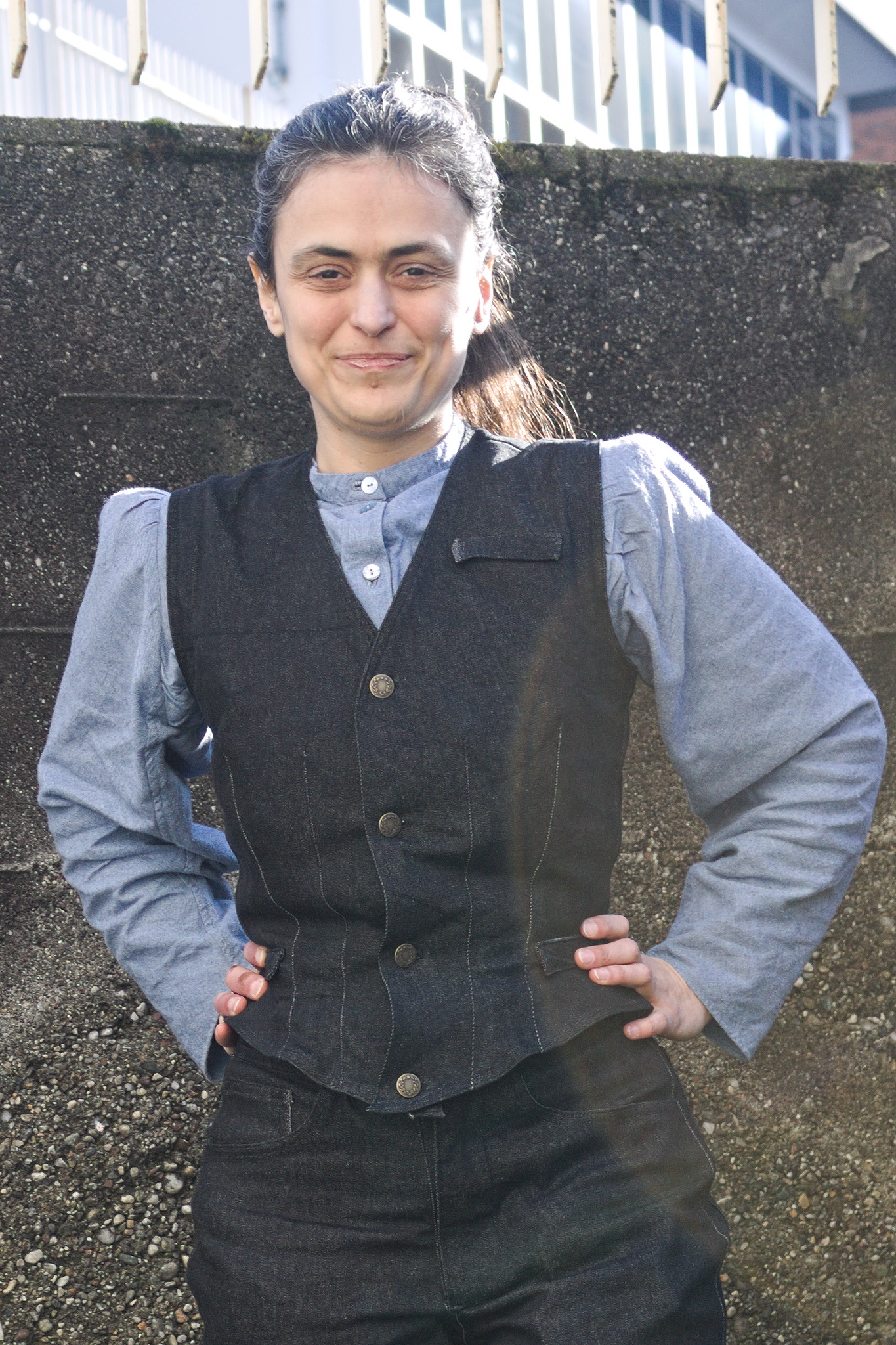 A woman wearing a single breasted waistcoat with double darts at the waist, two pocket flaps at the waist and one on the left upper breast. It has four jeans buttons.