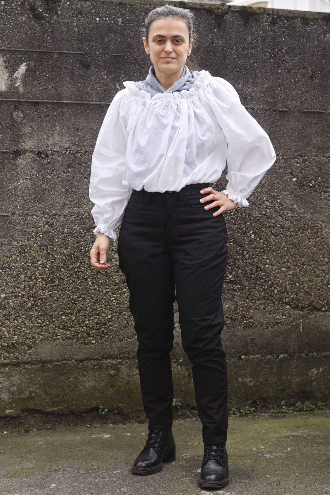 A woman wearing a white top with a wide neck with ruffles and puffy sleeves that are gathered at the cuff. The top is tucked in the trousers to gather the fullness at the waist.