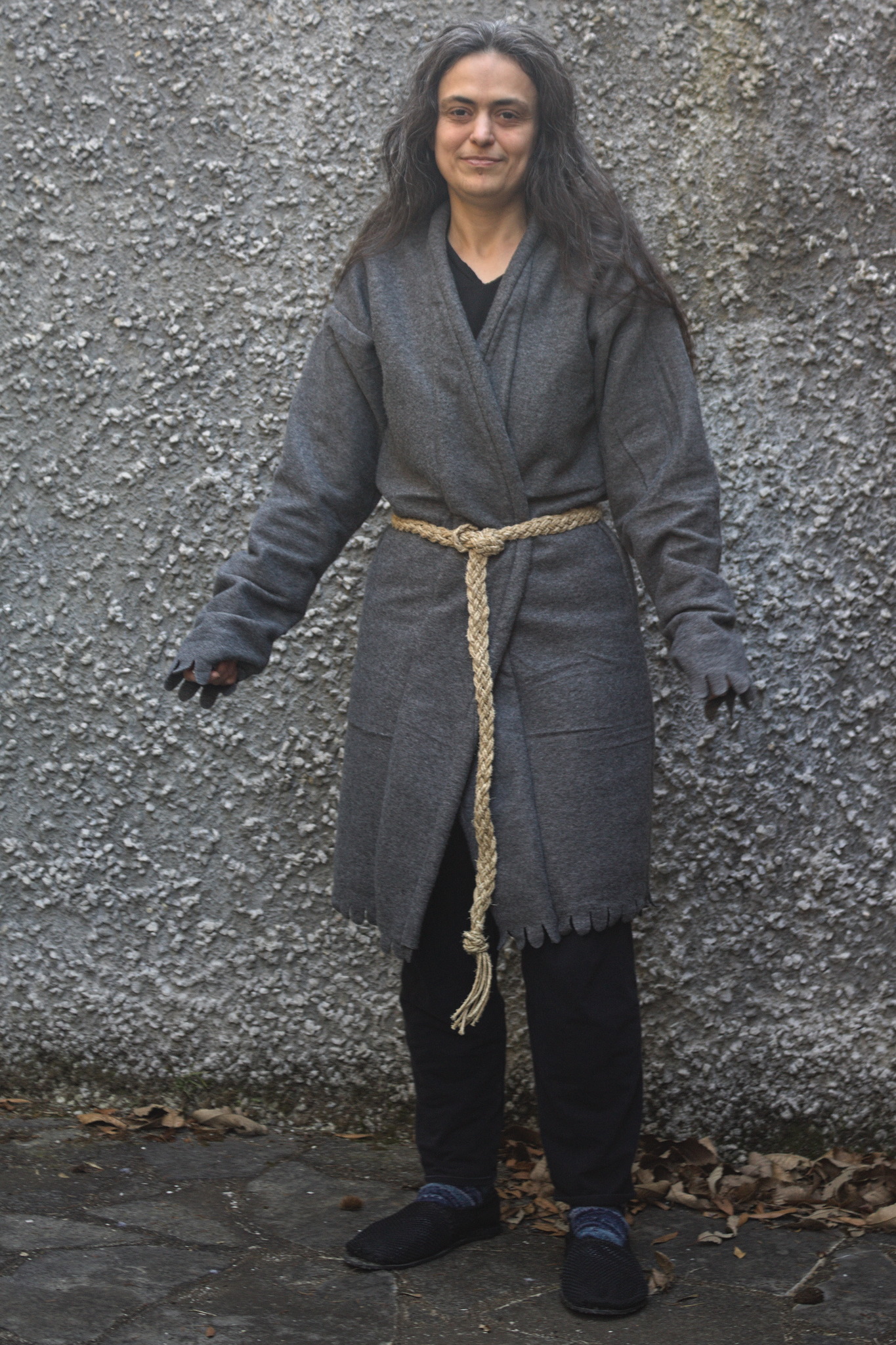 A woman with early morning hair wearing a knee-length grey
polar fleece jacket; it is closed at the waist with a twine belt
that makes the bound front edge go in a smooth curve between the
one side of the neck to the other side of the waist and then flare
back outwards on the hips.
The sleeves are long enough to go over the hands, and both them
and the hem are cut