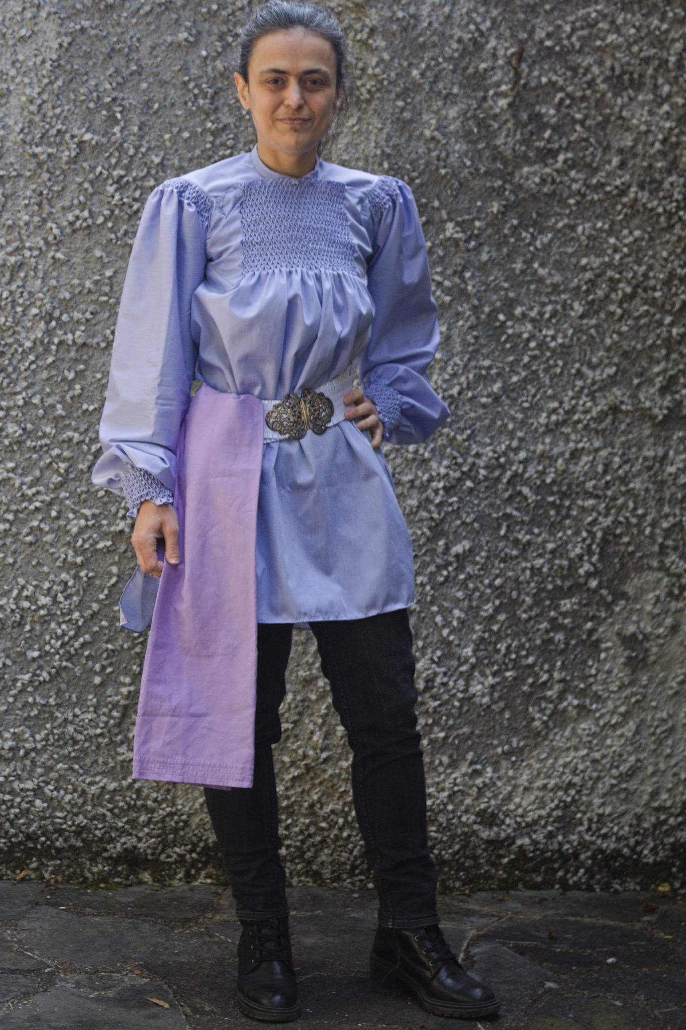 A woman wearing a purplish blue shirt with very wide sleeves,
gathered at the cuffs and shoulder with honeycombing, and also a
rectangle of honeycombing in the front between the neckline and
just above the bust.
The shirt is gathered at the waist with a wide belt, and an almost
lilac towel hangs from the belt.