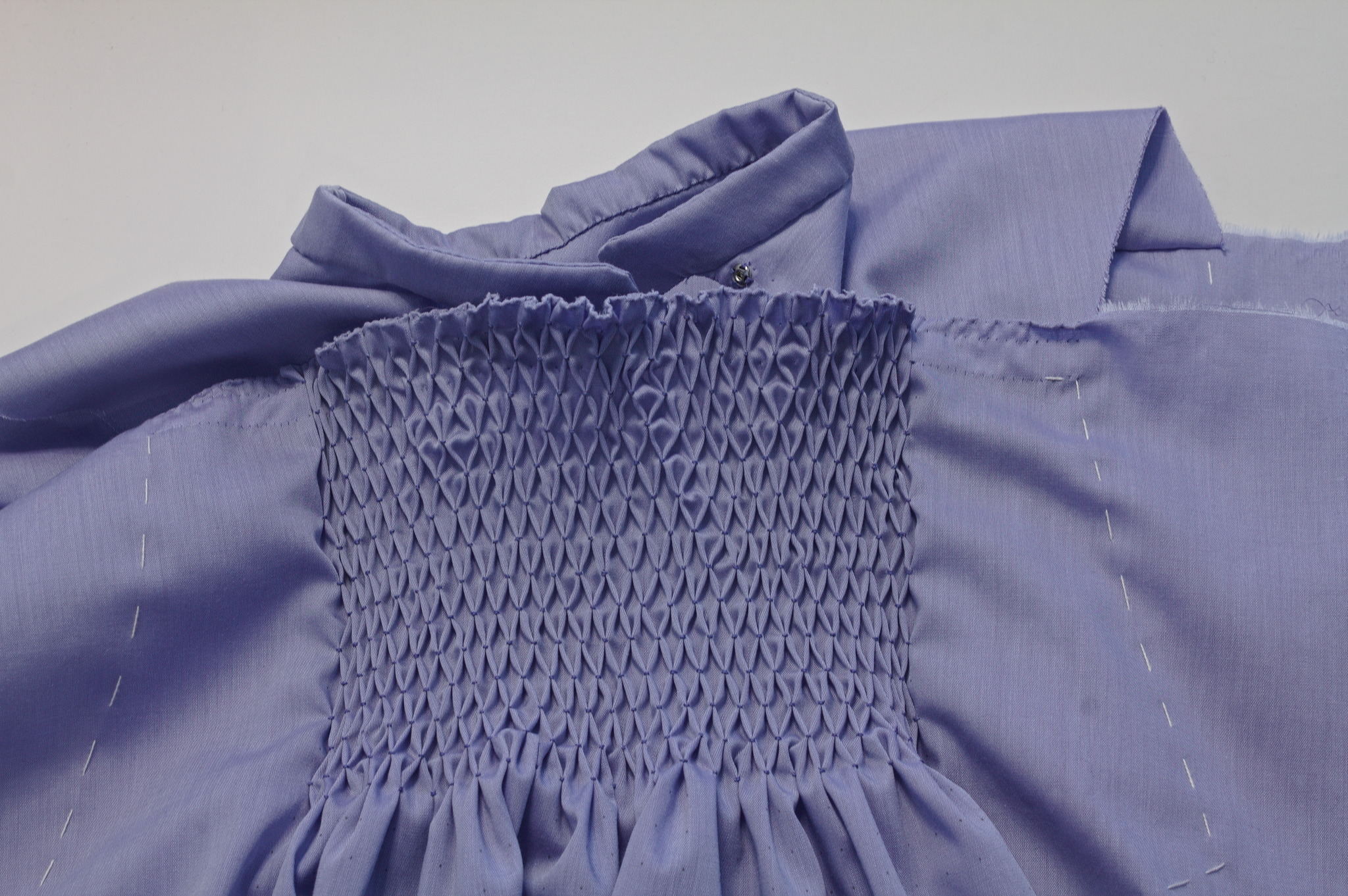 A piece of fabric with many rows of honeycombing laid on top of
the collar and yoke of the shirt; a metal snap peeks from behind
the piece of honeycombed fabric.

There are still basting lines for the armscyes.