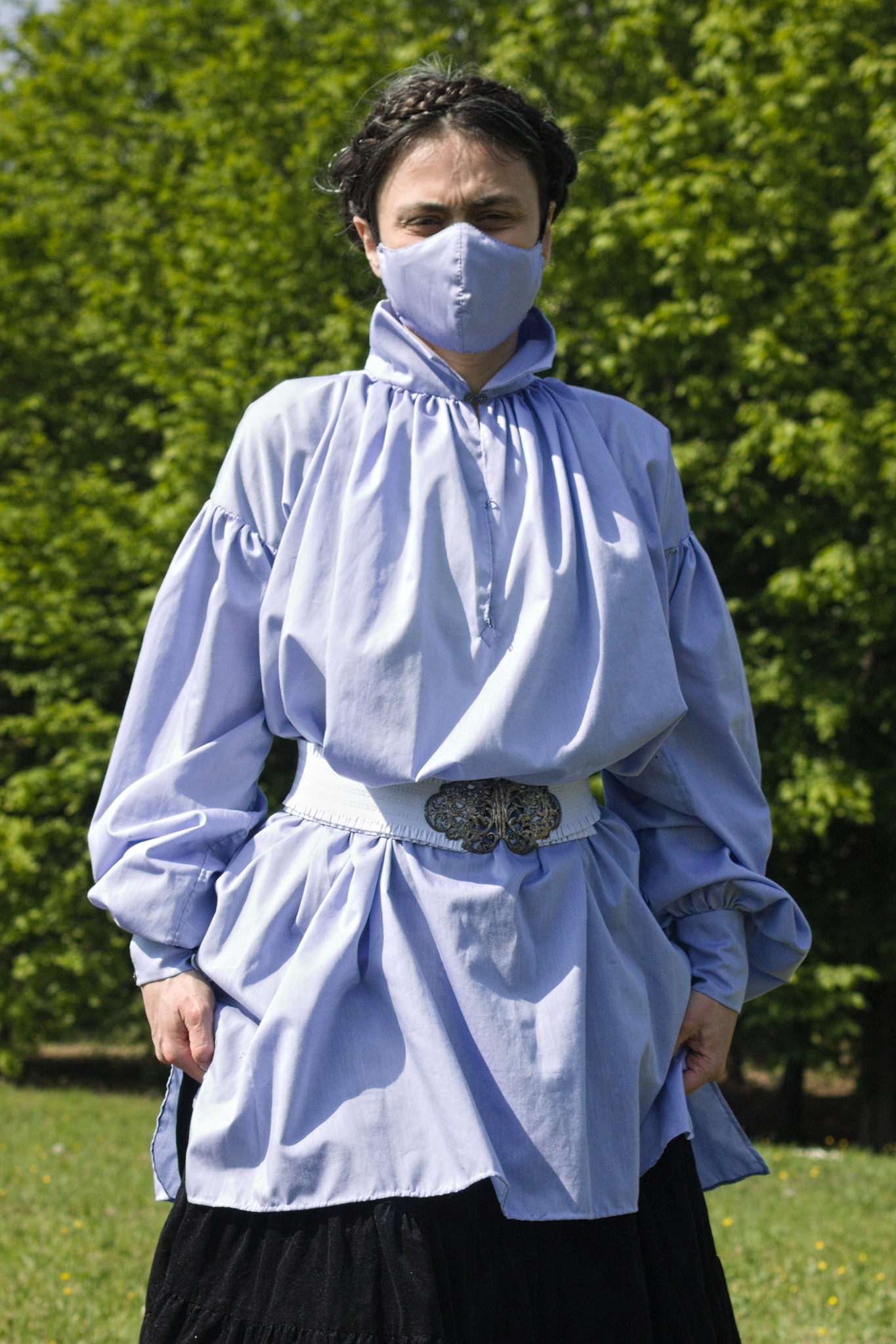 A woman wearing a shirt in the same fabric; this one has a slit
in the front, is gathered into a tall rectangular collar and has
dropped shoulders because it's cut from plain rectangles. The
sleeves are still huge, and gathered into tall cuffs.
It is worn belted (with the same wide white elastic belt used in
the previous picture) and the woman is wearing a matching fabric
mask, because the picture has been taken in 2021.