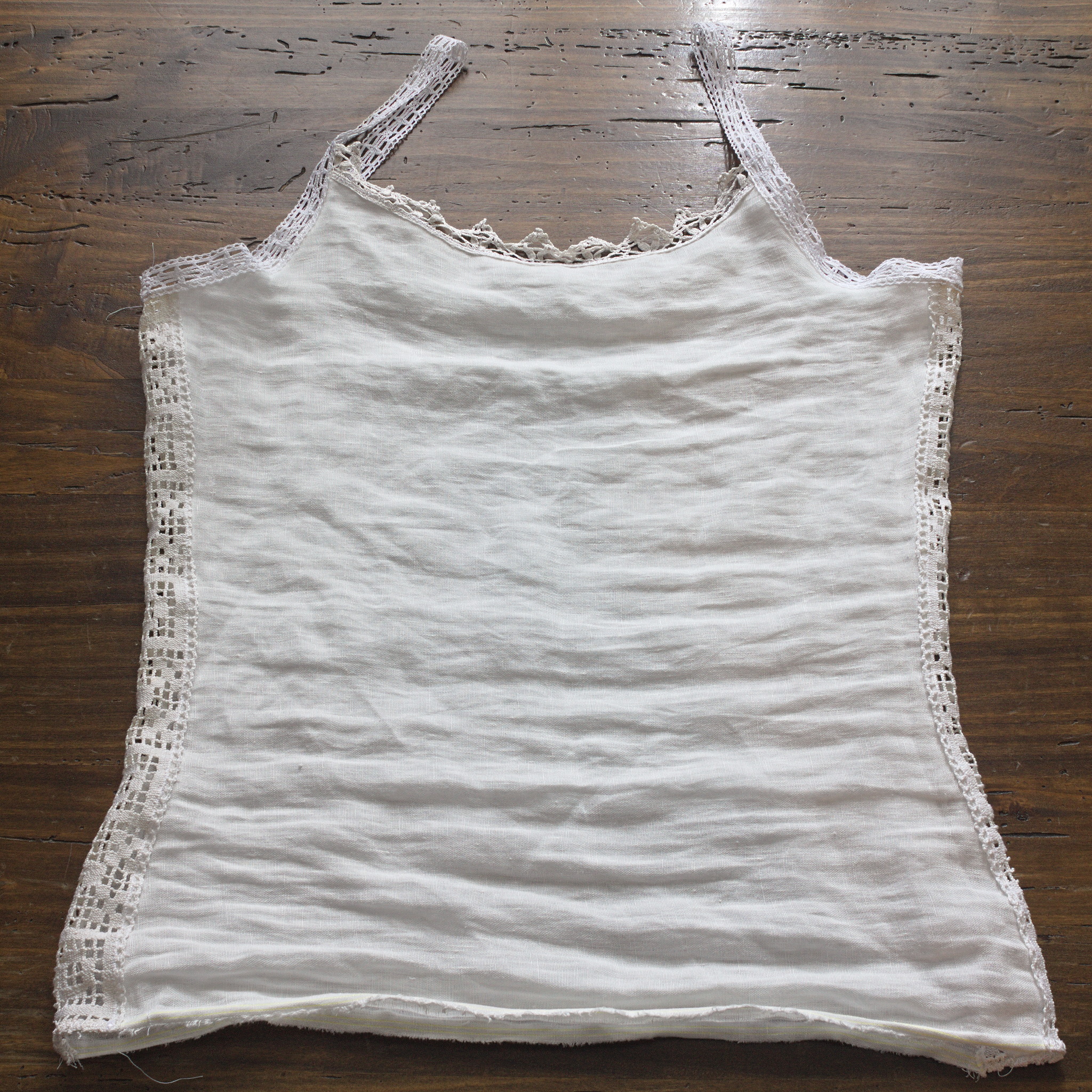 A camisole of white linen fabric; the sides have two vertical strips of filet cotton lace, about 5 cm wide, the top of the front is finished with another lace with triangular points and the straps are made with another insertion lace, about 2 cm wide.