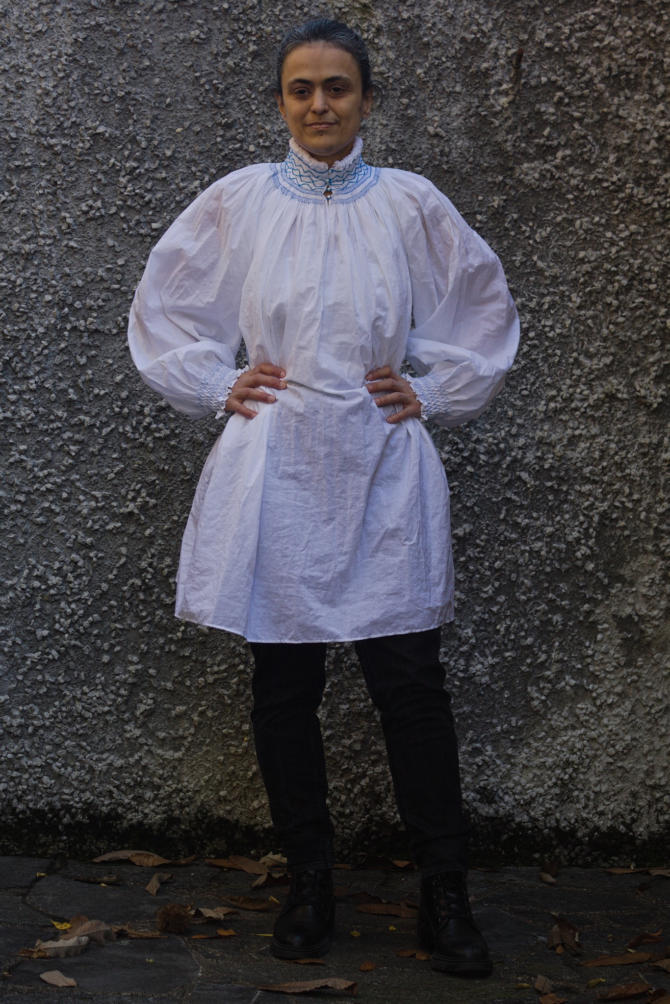 A woman wearing a white shirt with a tall, thick collar with
lines of blue embroidery, closed in the front with small buttons;
the sleeves are wide and billowing, gathered at the cuffs with
more blue embroidery. She's keeping her hands at the waist so that
the shirt, which reaches to mid thigh, doesn't look like a
shapeless tent from the neck down.