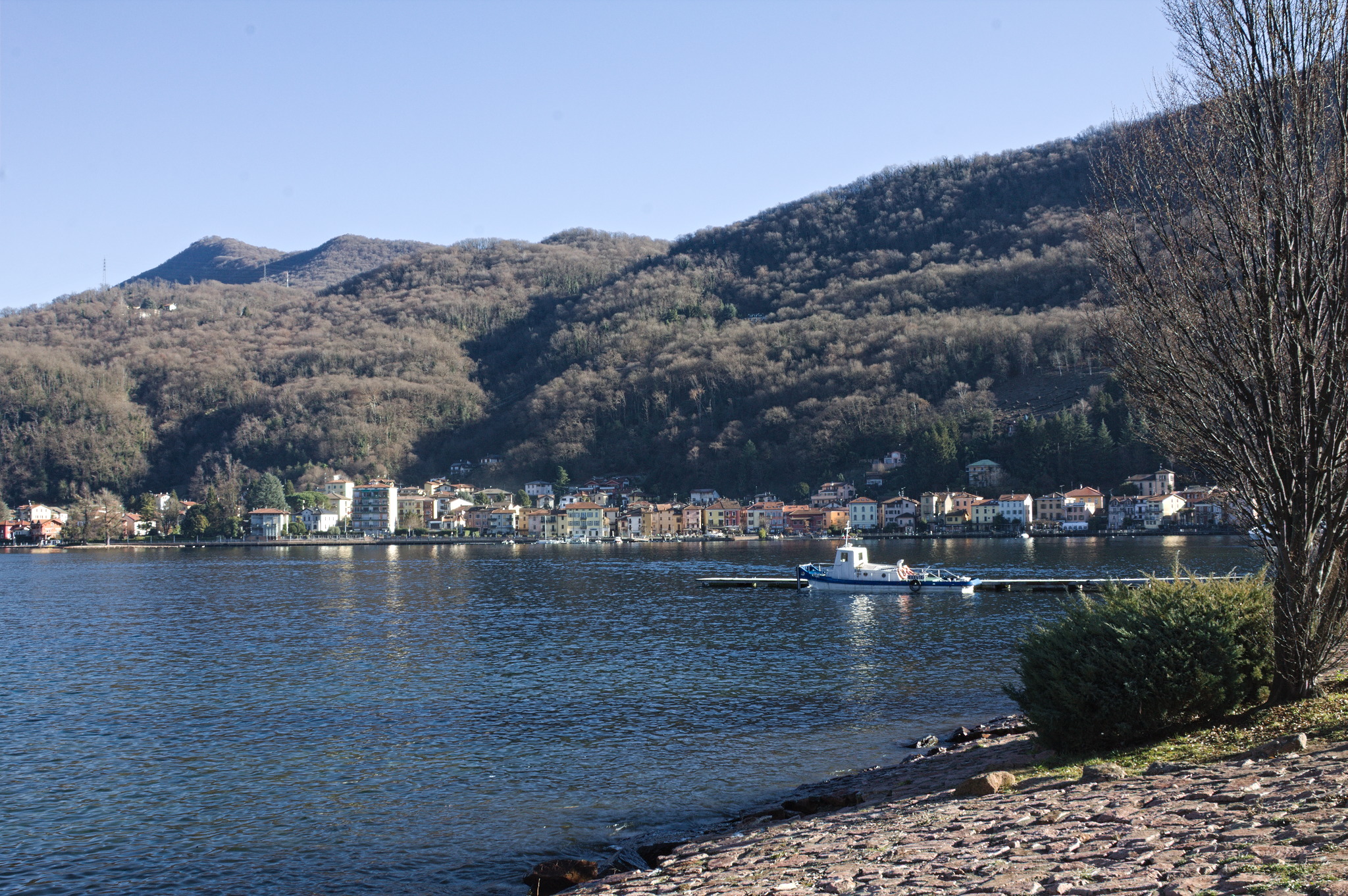 One side of Porto Ceresio along the lake: there is a small strip of houses, and the hills behind them are covered in woods. A boat is parked around the middle of the picture.