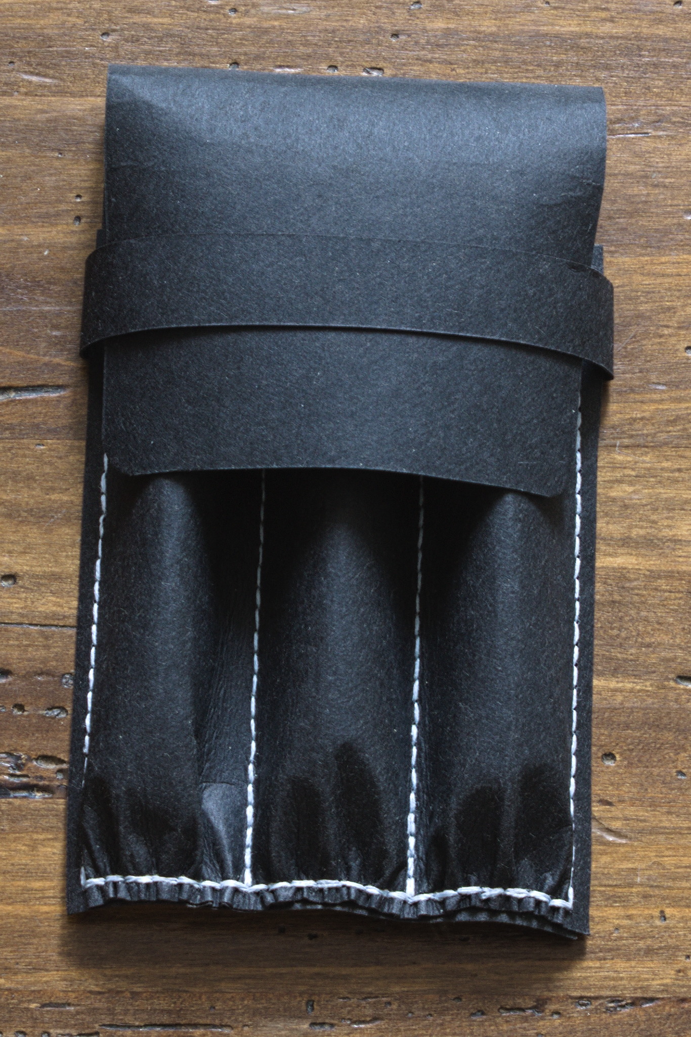 A pen case made of two pieces of a relatively stiff black material with a flat base and three separate channels on top, plus a flap covering everything and a band to keep the flap closed; there is visible light blue stitching all around the channels.