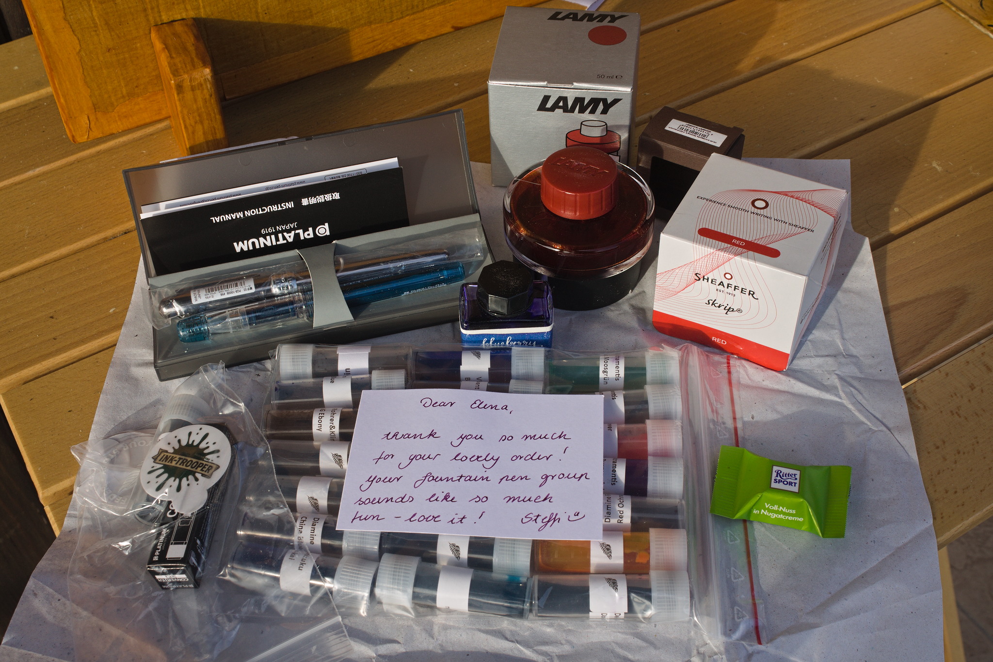 The contents of an order from a fountain pen shop, spread out
on a table: a couple of cheap pens, a couple bottles of ink, a
converter, a small ritter sport chocolate and a bag full of 5
ml vials with 2 ml of ink each, and a thank-you note from the
seller (Steffi).