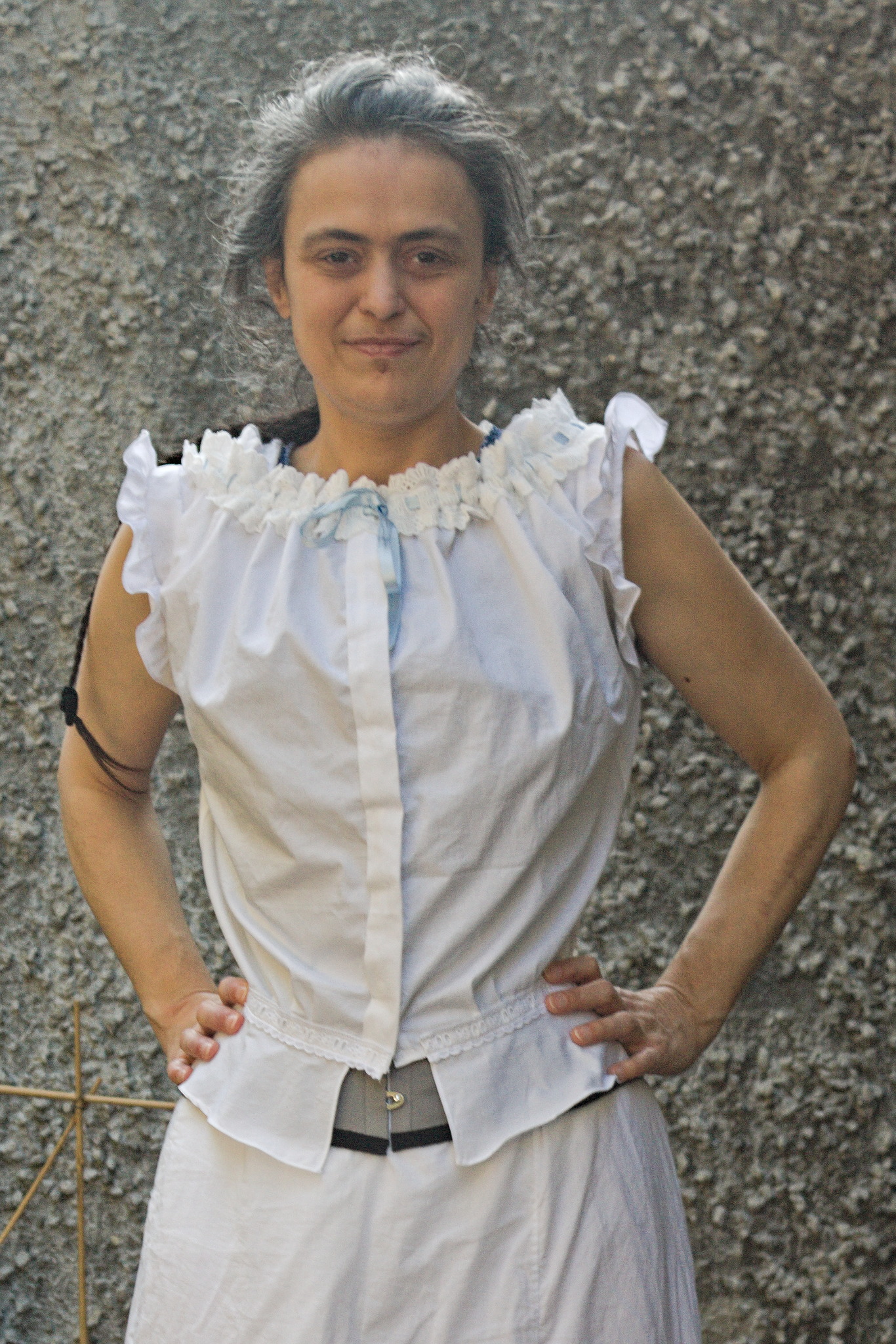 A woman wearing a sleeveless blouse in white fabric with a big
band of whitework embroidery gathered over a light blue ribbon at
the neckline, a box pleat at the front, another, smaller, band of
whitework embroidery at the waist, without a ribbon, and a short
peplum that doesn't reach the center front. Around the armscyes
there are small ruffles, giving even more volume at the top.
A bit of a grey corset peeks out from the center front, below the
waist.