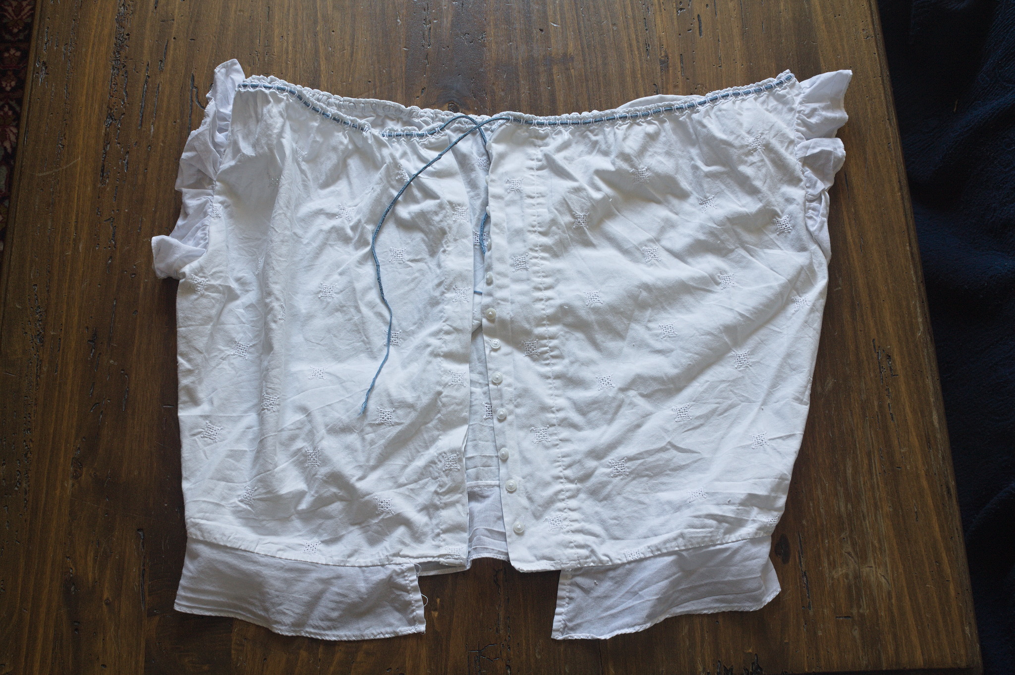 A sleeveless blouse in white fabric with machine whitework
embroidery; it has small ruffles around the armscyes and the
neckline is low and wide, with beading lace and a blue cord going
through it to gather it up.